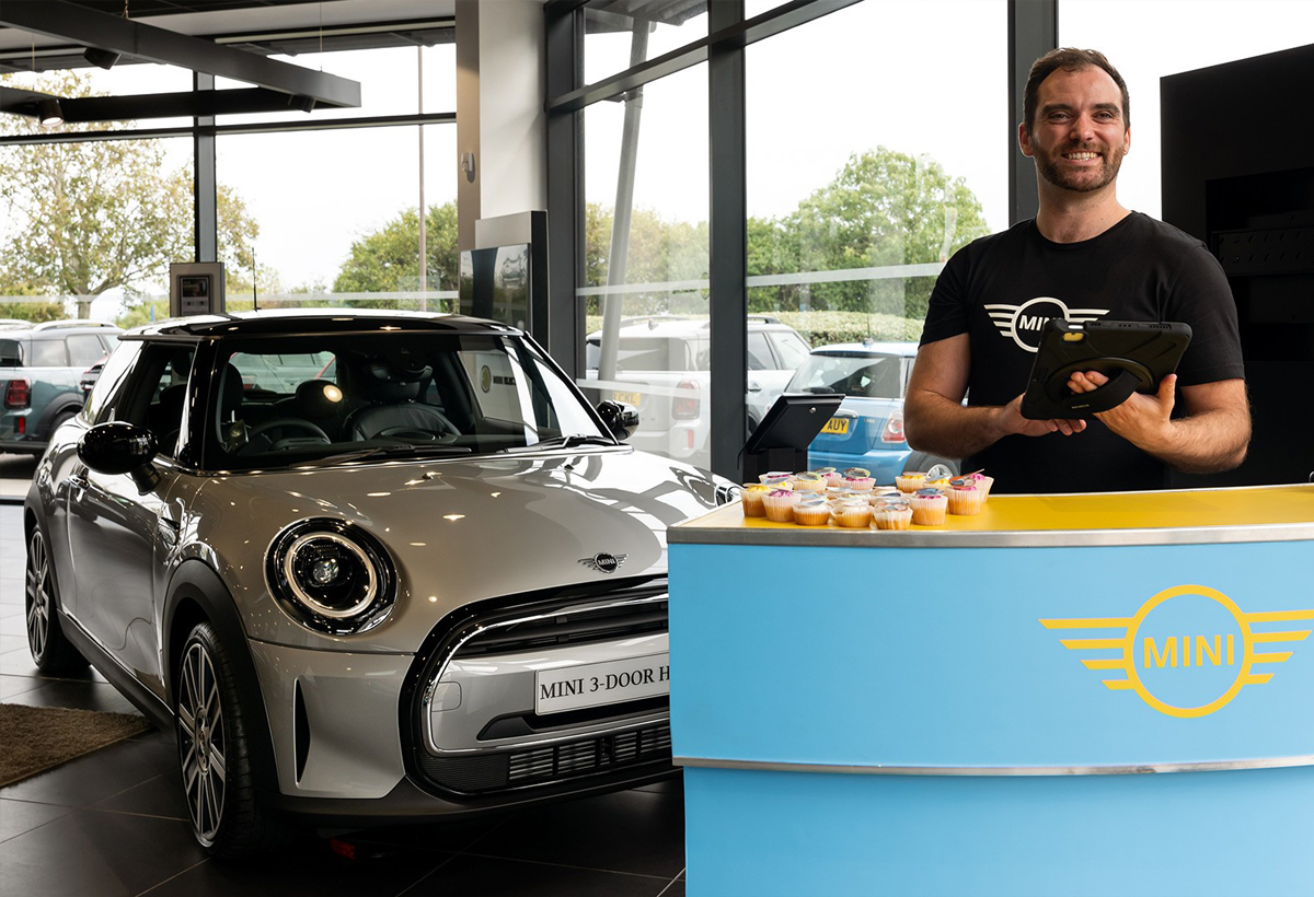 MINI 3-door hatchback car displayed in a showroom with a person behind a blue counter holding a tablet and cupcakes on the counter.